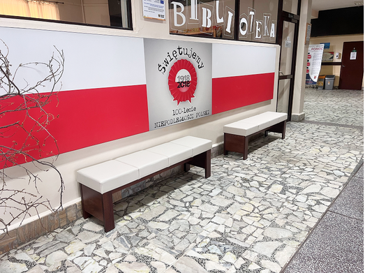 Ławki tapicerowane białą tkaniną, wykonane z litego drewna, ustawione na szkolnym korytarzu.